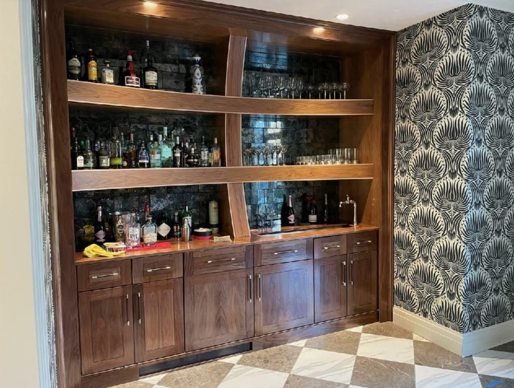 A built-in wooden bar with shelves holding bottles and glassware, set against a patterned wallpaper and checkered floor.