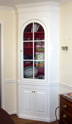 White corner cabinet with glass door, red interior shelves, and various dishes and glassware displayed inside.
