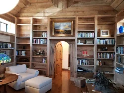 Wood-paneled library with built-in bookshelves, an armchair, a round table, and a painting above a wooden arched door.