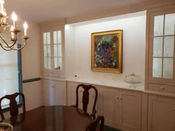 Dining room with a wooden table, glass-front cabinets, and a colorful framed painting on a built-in white wall.