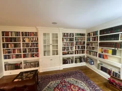 A home library with built-in white bookshelves filled with books, a glass cabinet, and a patterned rug on wooden floor.