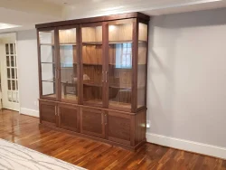 A large wooden display cabinet with glass doors and shelves stands against a light gray wall on a polished wood floor.