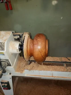 A wooden bowl with visible cracks is being turned on a lathe, surrounded by wood shavings in a workshop.