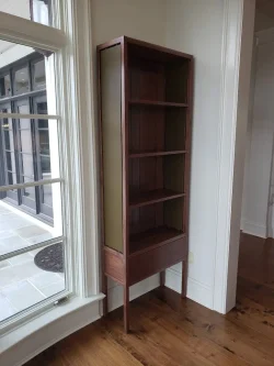 A tall, narrow wooden bookshelf with four shelves stands in the corner near a large window on a hardwood floor.