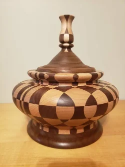 A wooden lidded bowl with geometric patterns in light and dark wood, sitting on a wooden surface.