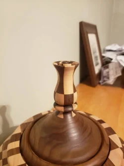 A wooden vase-shaped handle on a round, striped wooden lid, with papers and a framed photo in the background.