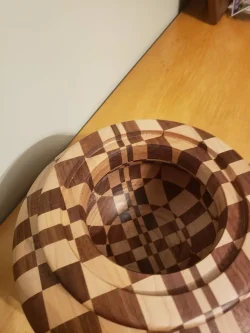 A wooden bowl with a checkered pattern sits on a wooden surface near a light-colored wall.
