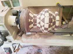 A wooden vase with a geometric checkerboard pattern is being shaped on a lathe in a woodworking shop.