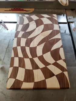 Rectangular wooden cutting board with an abstract wavy pattern of alternating light and dark wood pieces.