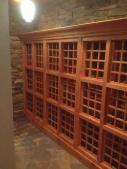 Wooden wine racks with grid-style compartments line a stone wall in a dimly lit room with tiled flooring.