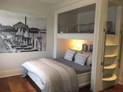 A bedroom with a double bed, loft space above, spiral stairs, and a large sailboat photo on the wall.