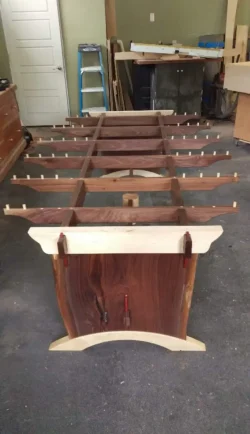 A large wooden frame with multiple dowels and clamps sits in a workshop, surrounded by tools and wood pieces.