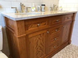 Wooden bathroom vanity with marble countertop, dual sinks, silver faucets, soap dispensers, and tiled backsplash.