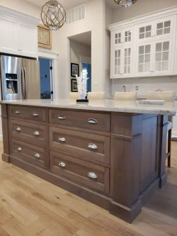 Brown kitchen island with silver handles is centered on a wood floor, with white cabinets and chairs in the background.