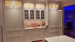 Well-lit kitchen with white cabinets, glass-front uppers, marble countertop, and cutting board on backsplash.