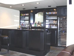 Modern home bar with black cabinetry, shelves of glassware and liquor bottles, and a built-in wine cooler.
