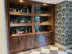 A wooden home bar with shelves holding bottles and glasses, set against a dark brick wall and patterned wallpaper.
