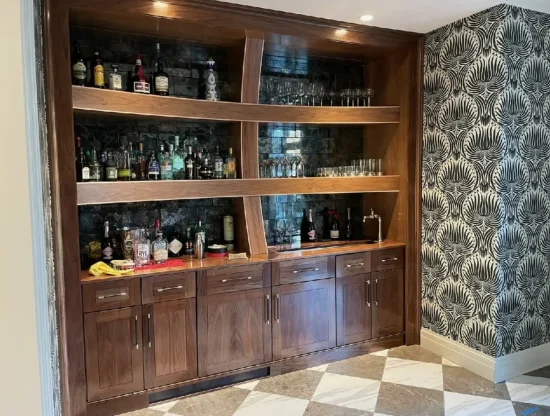 A wooden home bar with shelves holding bottles and glasses, set against a dark brick wall and patterned wallpaper.