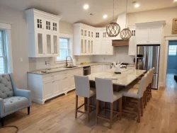 Modern white kitchen with an island, beige chairs, stainless steel appliances, and pendant lights.