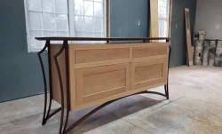 A wooden sideboard with arched legs and light wood panels stands in an unfinished room with large windows.