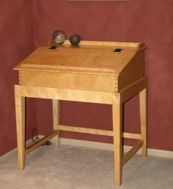 A wooden writing desk with a slanted lid, two decorative spheres on top, against a red textured wall on a carpeted floor.