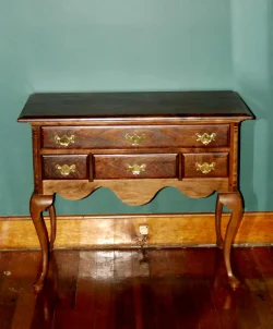 A small wooden side table with three drawers and brass handles stands against a teal wall on a hardwood floor.